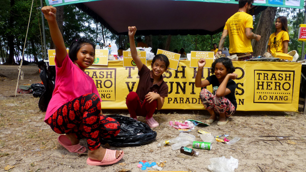 Three girls from thailand are crouching on the floor putting an arm in the air triumphently. They are smiling at the camera in front of a trash hero Ao Nang sign